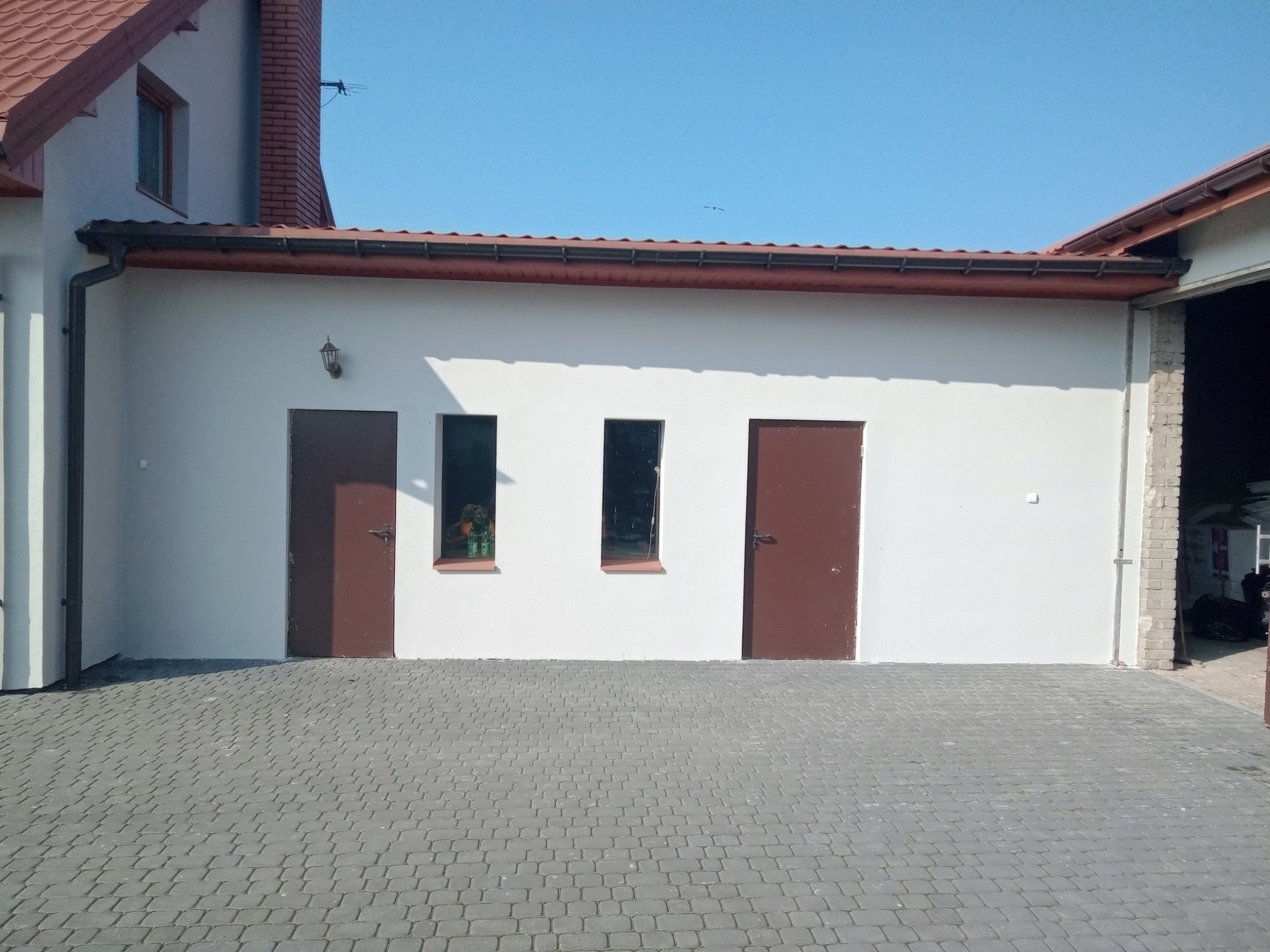 Świeżo otynkowana, jasna elewacja budynku gospodarczego z dwoma brązowymi drzwiami i dwoma małymi oknami, widok z poziomu kostki brukowej.