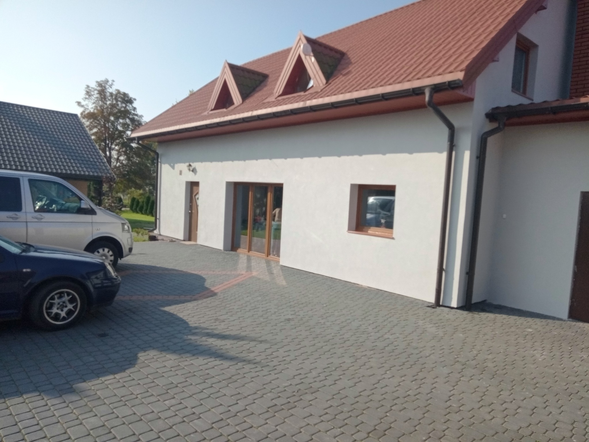 Jasna elewacja domu z brązowym dachem i stolarką okienną, widoczny szary bruk i zaparkowane samochody w słoneczny dzień.