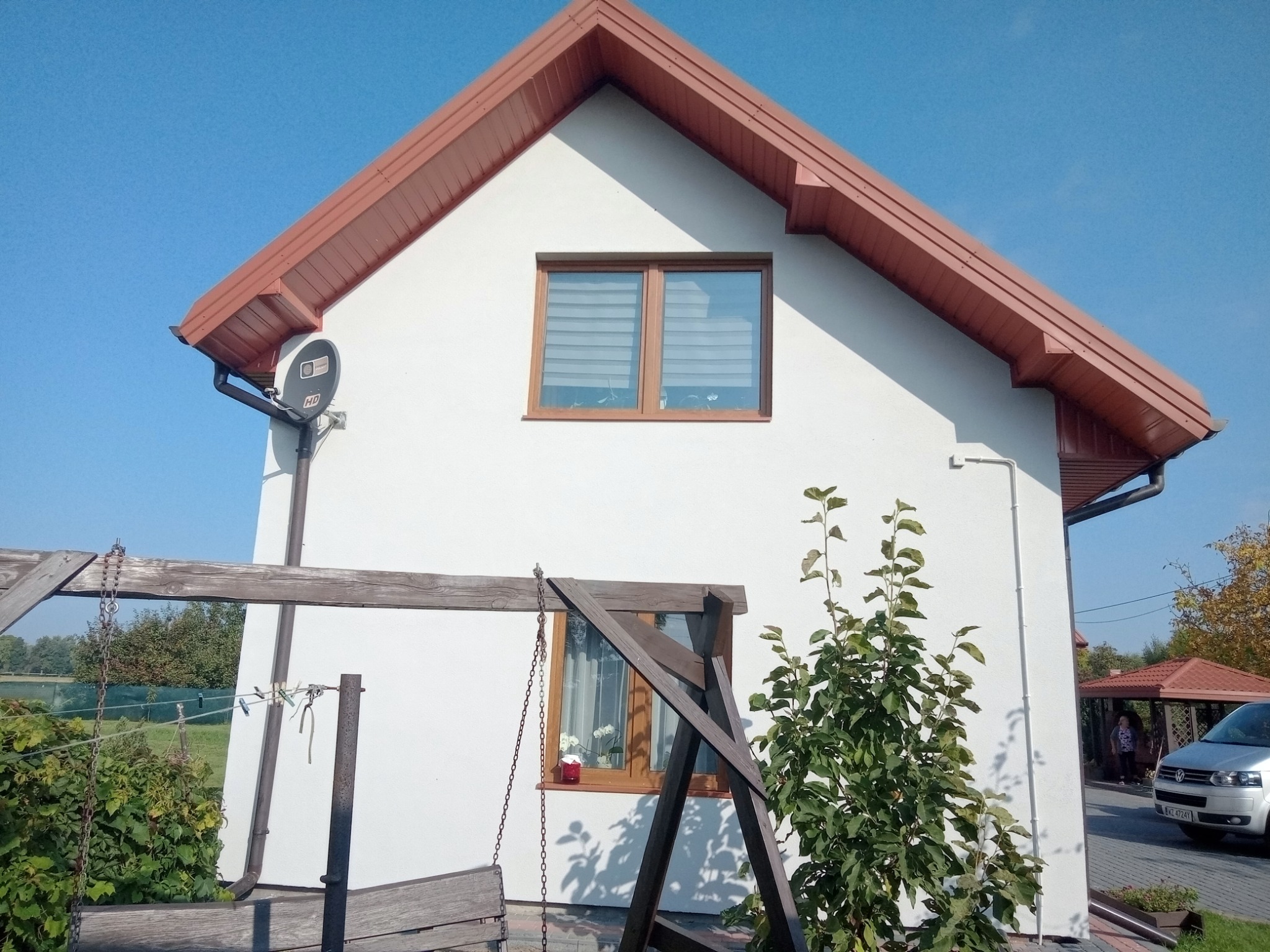 Dom jednorodzinny z białą elewacją, brązowym dachem, oknami z żaluzjami i drewnianą huśtawką na pierwszym planie. Z boku widoczny fragment ogrodu i samochodu.