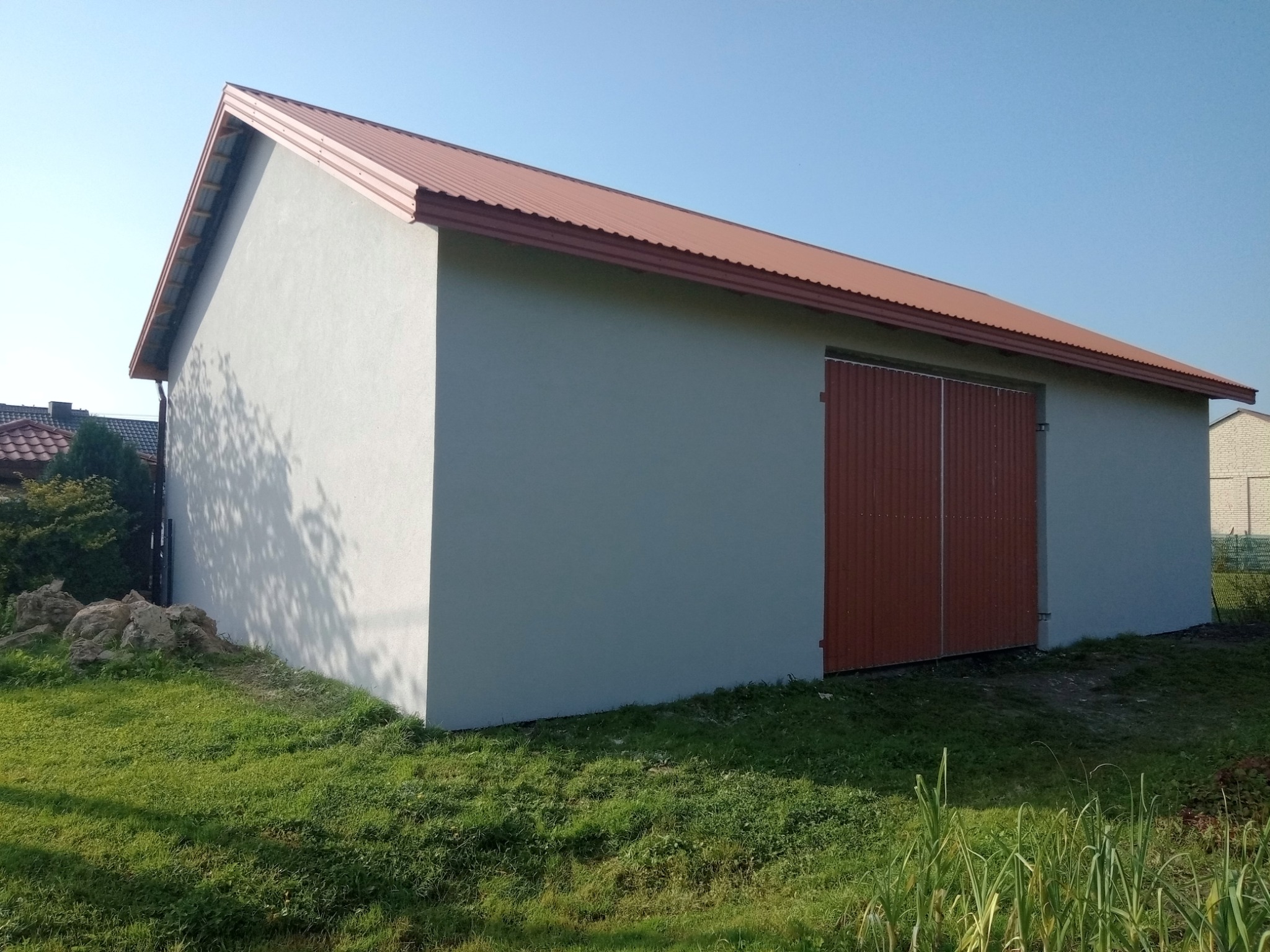 Świeżo otynkowany budynek gospodarczy z brązowymi drzwiami i dachem, widoczny z poziomu trawnika w słoneczny dzień.