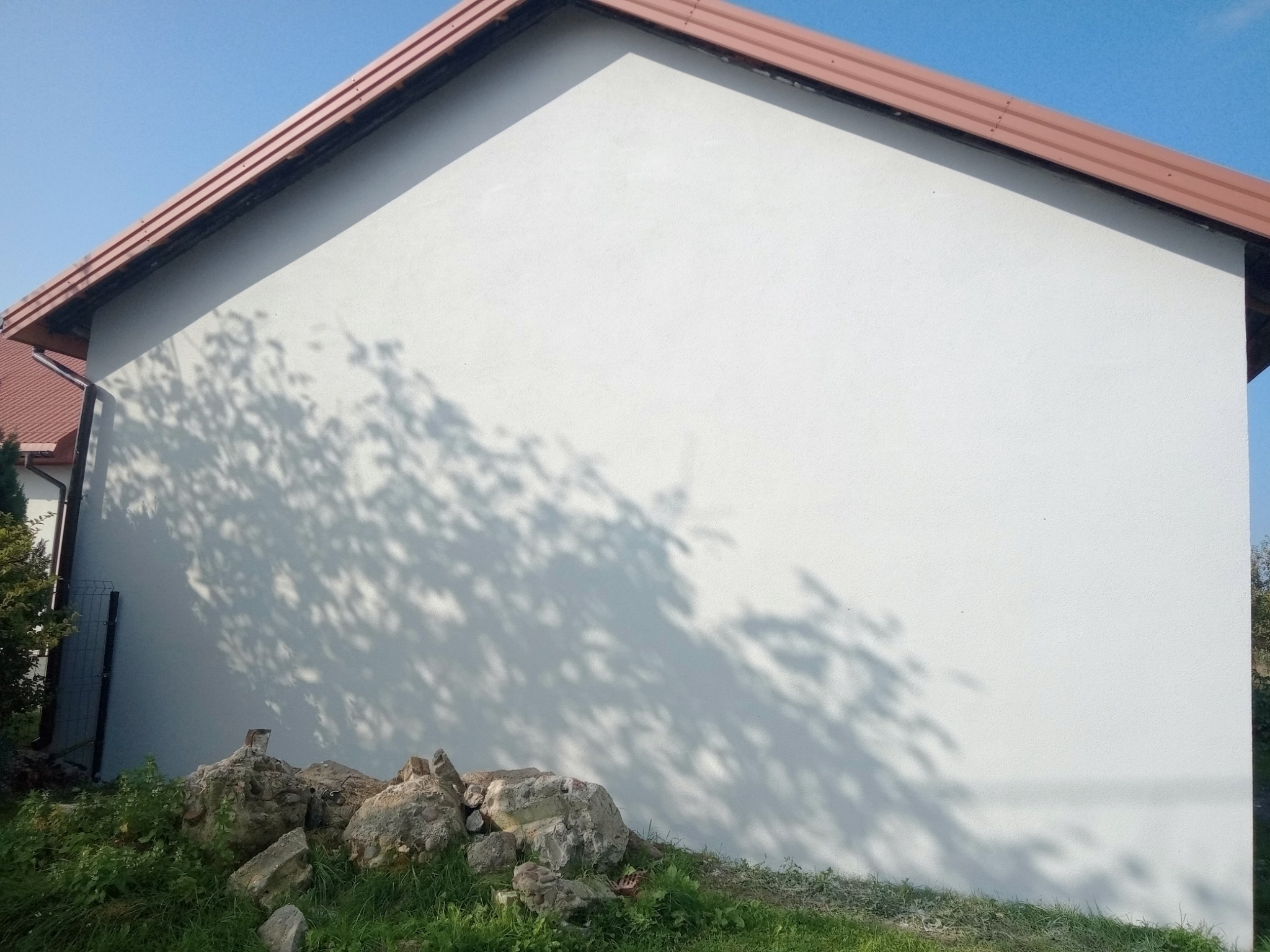 Świeżo otynkowana ściana budynku z brązowym dachem, na której widoczne są cienie drzew, a u dołu leżą gruz i kamienie.