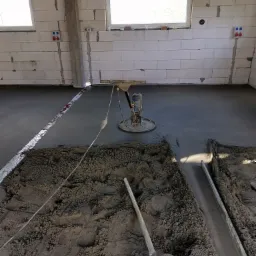 Wnętrze w trakcie wylewania posadzki, widoczna zacieraczka mechaniczna, świeżo wylany beton oraz pryzma mokrego piasku z łopatą i aluminiową łatą w tle surowych ścian z betonu komórkowego.