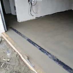 Świeżo wylana, gładka posadzka jastrychowa z widocznymi łatami poziomującymi i narzędziami murarskimi w surowym wnętrzu budynku.