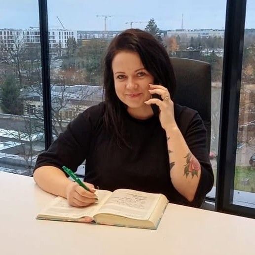 Kobieta w czarnej bluzce siedzi przy biurku, rozmawia przez telefon i notuje w otwartej książce, na ramieniu widoczny tatuaż, za nią duże okno z widokiem na miasto.