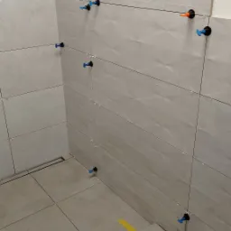 Świeżo ułożone, szare płytki w łazience, z systemem poziomowania płytek (klipsy i kliny) w kolorach niebieskim i pomarańczowym, oraz z żółtą taśmą malarską zabezpieczającą krawędzie.