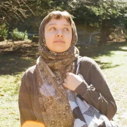 Portret kobiety z chustą na głowie, stojącej na trawniku z drzewami w tle, z torbą na ramieniu i spojrzeniem skierowanym w górę.