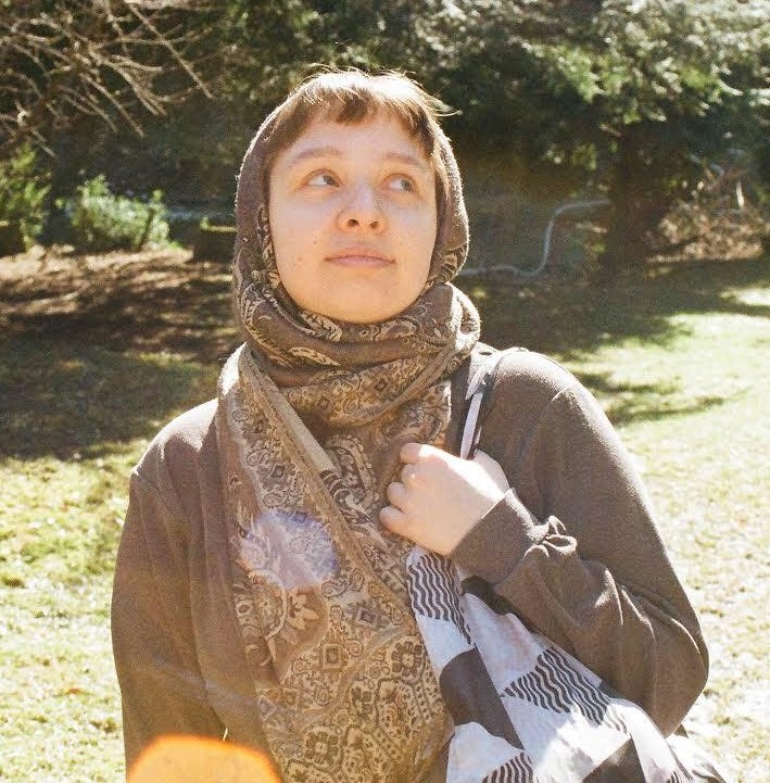 Portret kobiety z chustą na głowie, stojącej na trawniku z drzewami w tle, z torbą na ramieniu i spojrzeniem skierowanym w górę.