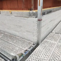 Wznoszenie ściany z białych bloczków na budowie domu, widoczne rusztowanie, izolacja termiczna i fragment ściany z czerwonej cegły w tle.