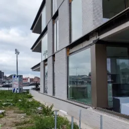 Surowy budynek z szarej cegły z dużymi oknami w trakcie budowy, widoczne rusztowanie, niezabezpieczone kable i gruz na ziemi.