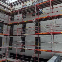 Wielopiętrowy budynek w trakcie budowy, otoczony rusztowaniami z widocznymi elementami konstrukcyjnymi i narzędziami na pierwszym planie, pochmurne niebo w tle.