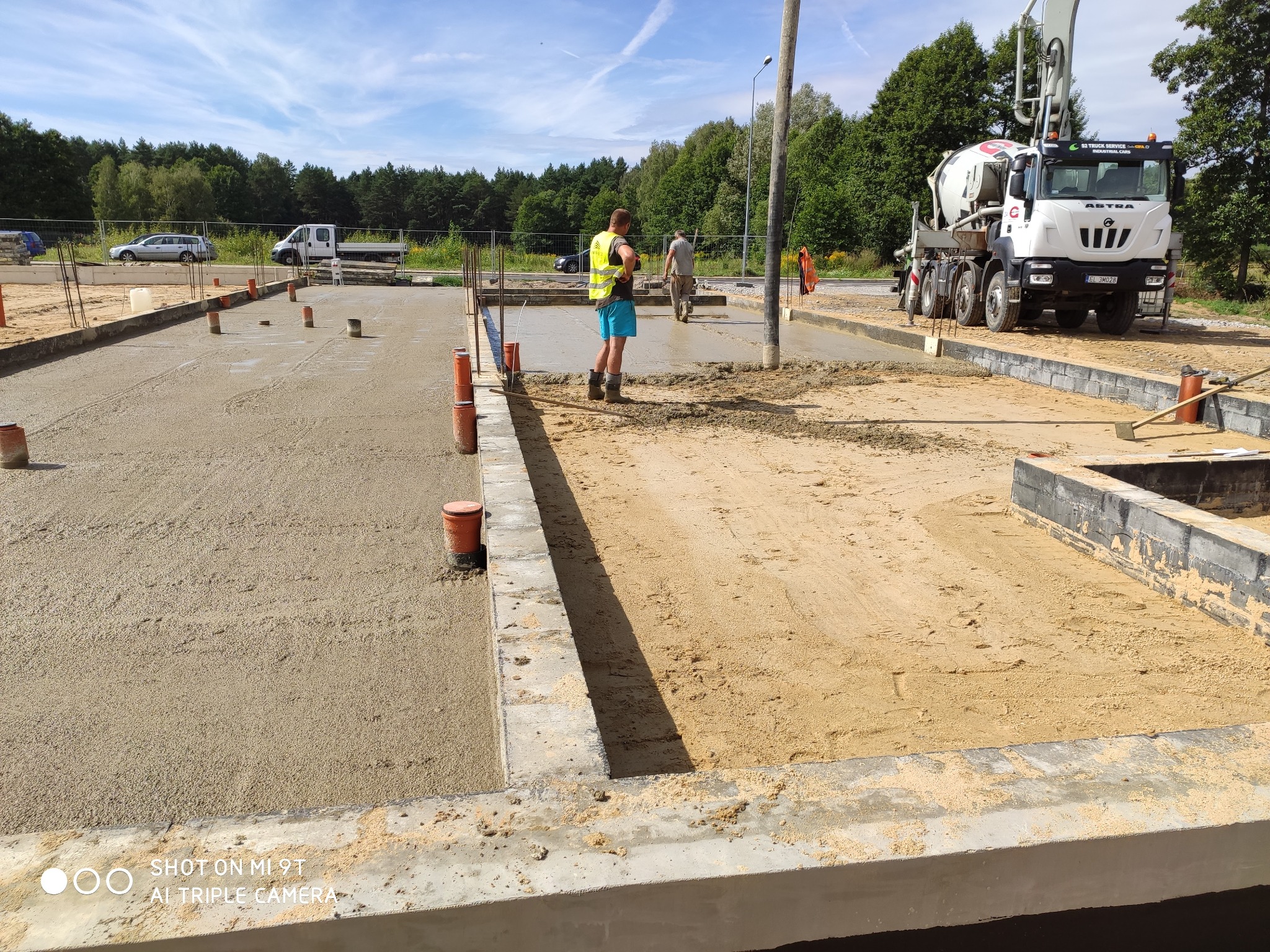 Wylewanie betonu na przygotowany fundament pod budowę domu, widoczny betonowóz i pracownicy budowlani w odblaskowych kamizelkach, na części powierzchni wylany beton, a na części piasek.
