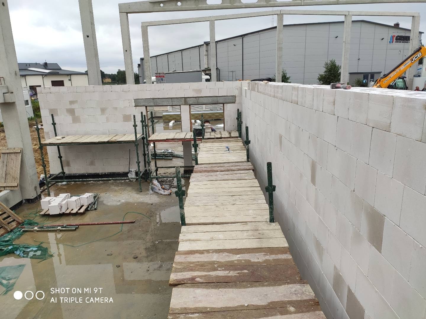 Wznoszenie ścian budynku z bloczków betonu komórkowego, widoczne rusztowania i drewniana kładka robocza, na zewnątrz plac budowy z widocznym budynkiem przemysłowym w tle.