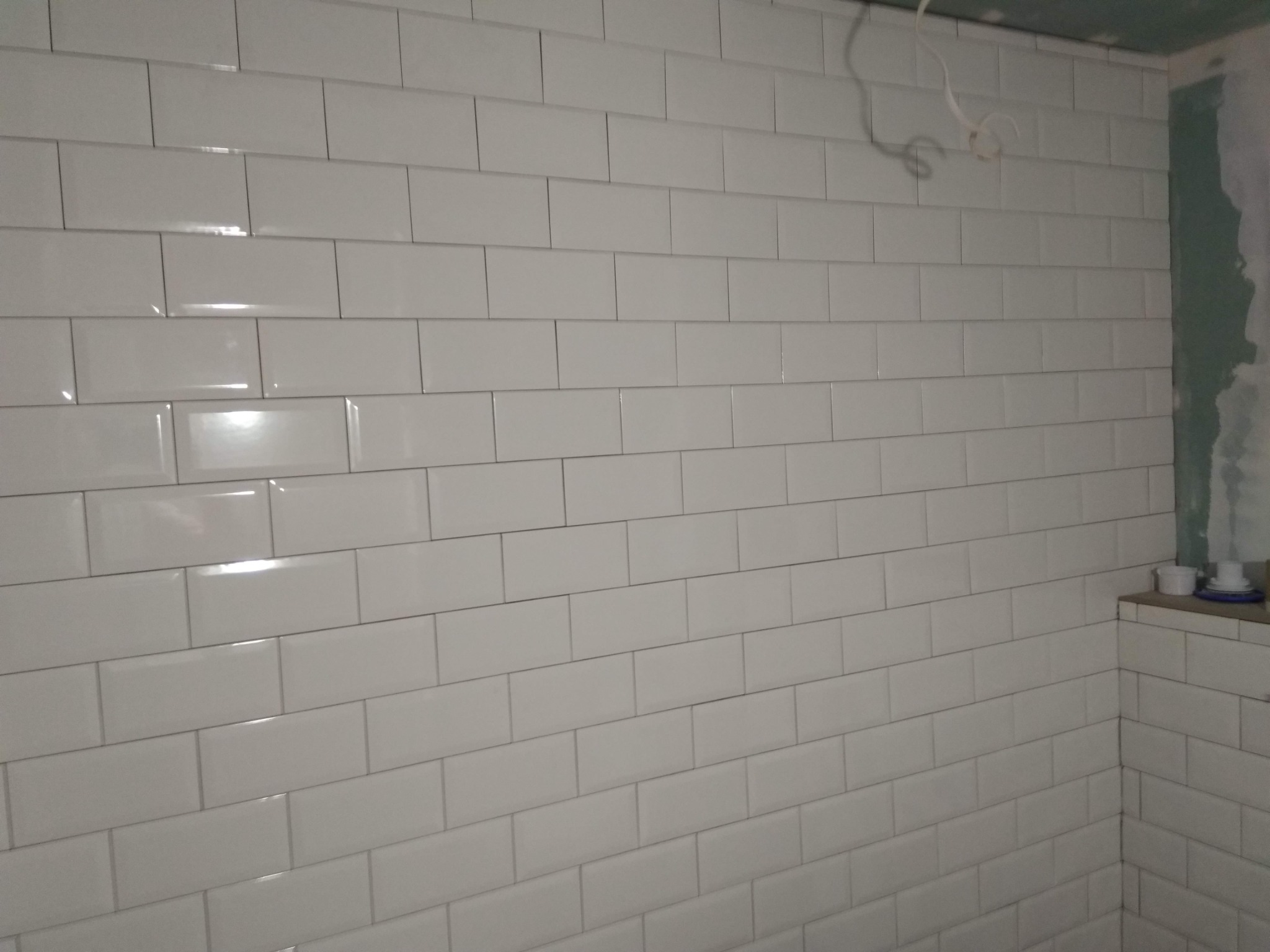 Ściana w trakcie układania białych, prostokątnych płytek ceramicznych z ciemną fugą; widoczne niedokończone wykończenia i wystające przewody elektryczne.