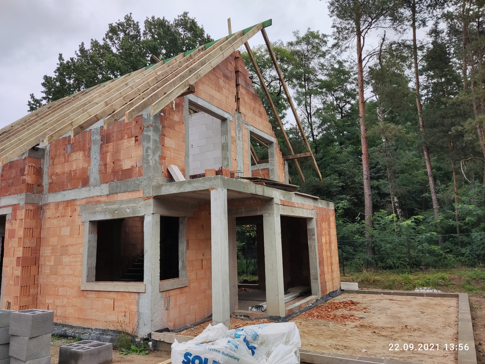 Surowy stan domu z czerwonej cegły z widocznym drewnianym szkieletem dachu na tle zieleni drzew, worki z zaprawą budowlaną leżące obok.