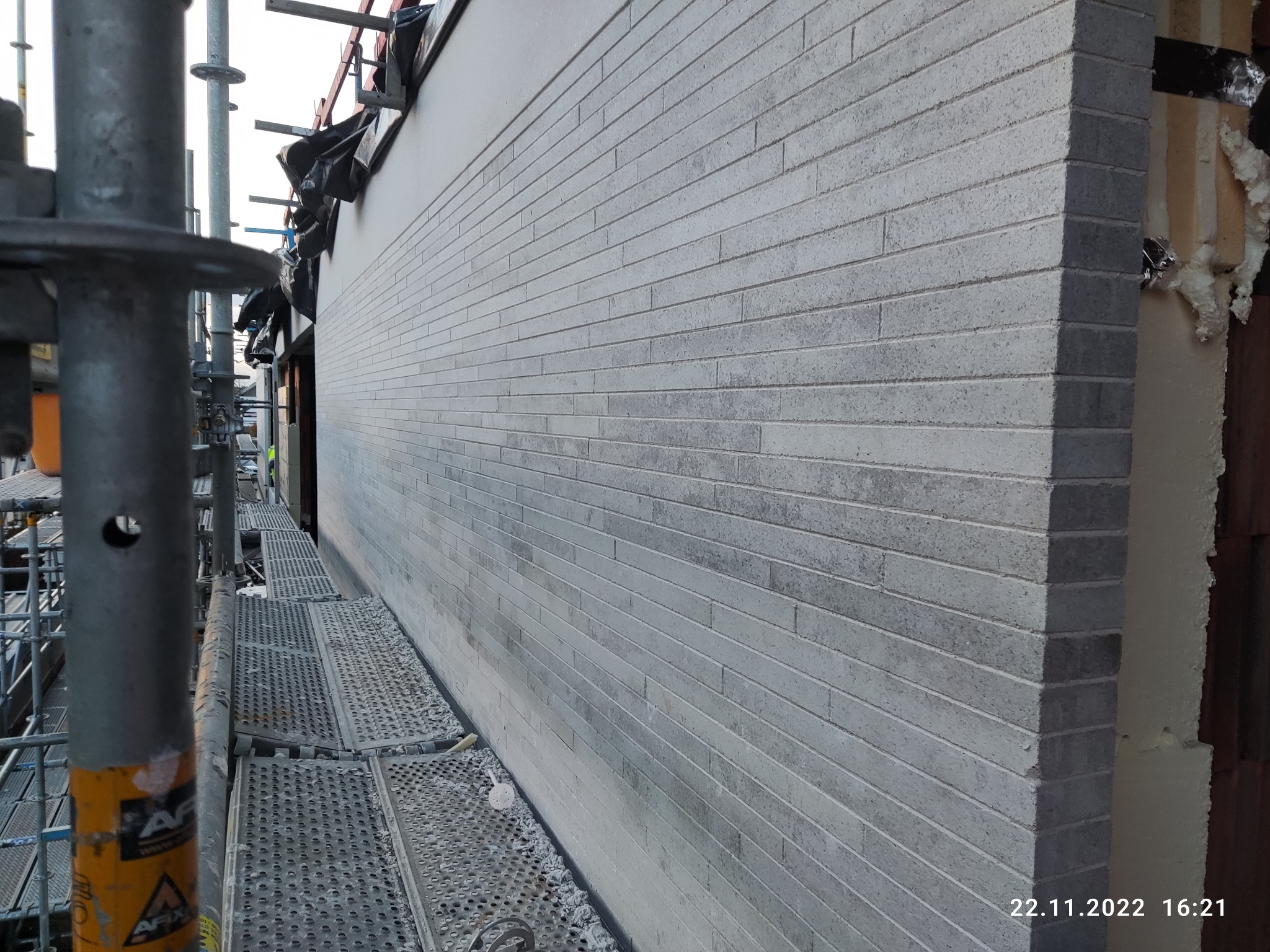 Ściana budynku w trakcie budowy, wykończona szarą cegłą licową, widoczne rusztowania i materiały izolacyjne.