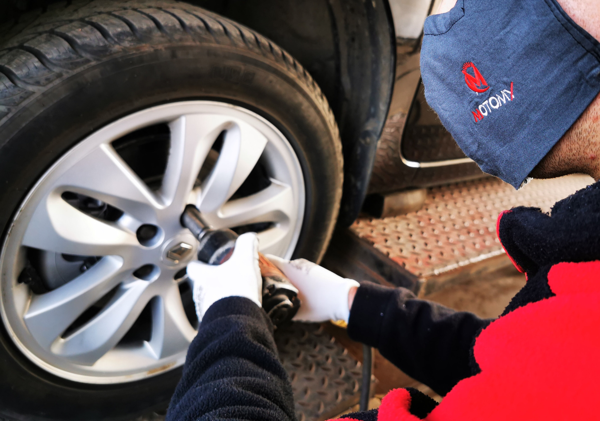 Pracownik w masce z logo firmy odkręca koło samochodu za pomocą klucza pneumatycznego, widoczne białe rękawice ochronne.