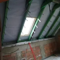 Poddasze w trakcie adaptacji: widoczne zielone krokwie, szara wełna mineralna między nimi, okno dachowe i fragment ściany z czerwonej cegły.