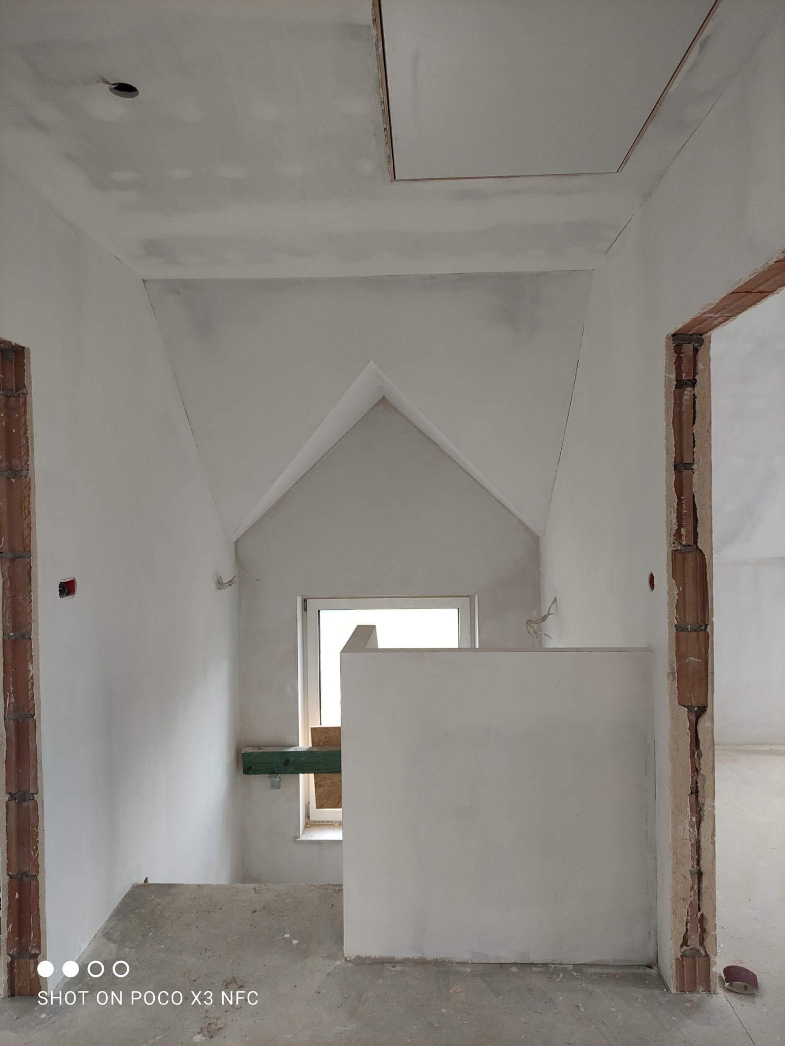 Poddasze w trakcie wykańczania: widoczne ściany i sufit po szpachlowaniu, z widocznymi śladami po łataniu, przygotowane do dalszych prac wykończeniowych, z oknem w tle i surowymi framugami drzwi.