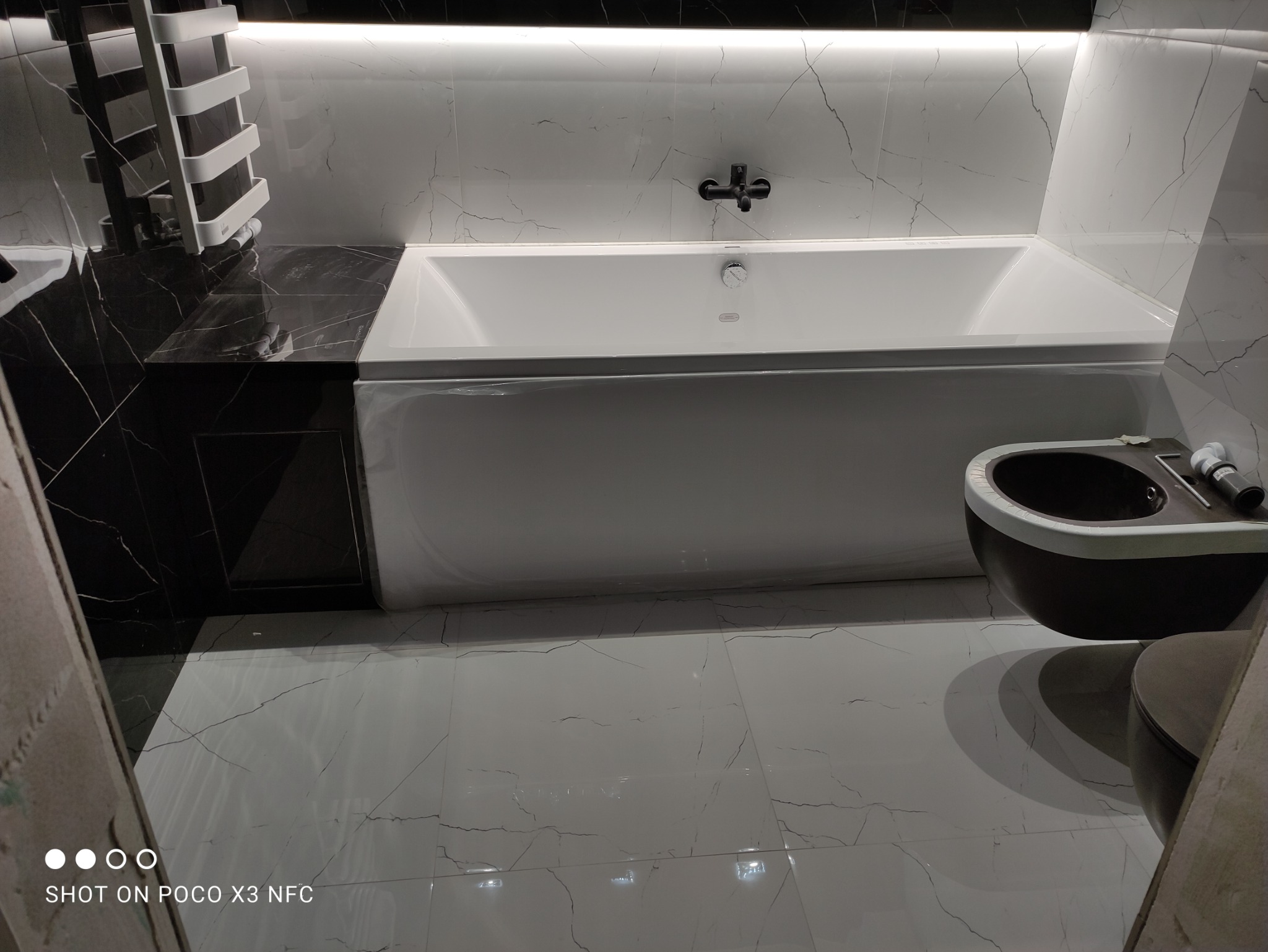 Nowoczesna łazienka z białą wanną, czarnym blatem z marmuru, biało-szarymi płytkami imitującymi marmur i czarnym bidetem, oświetlona listwą LED pod sufitem.