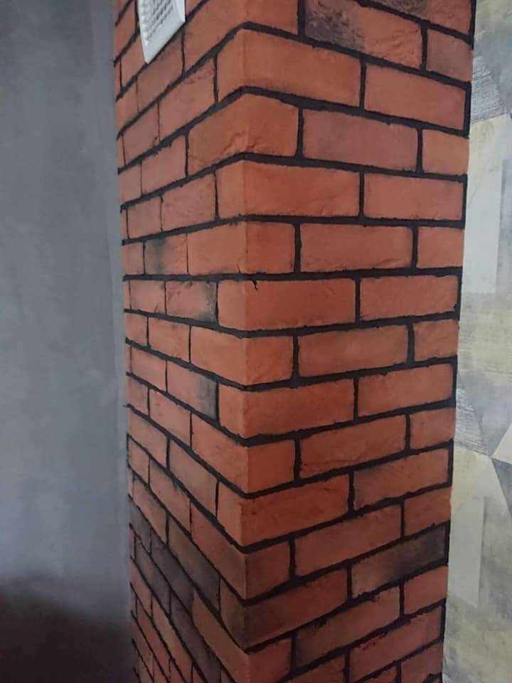 Dekoracyjny narożnik ściany wykończony czerwoną cegłą klinkierową z czarną fugą, widoczna kratka wentylacyjna u góry, na tle szarej ściany i wzorzystej tapety.