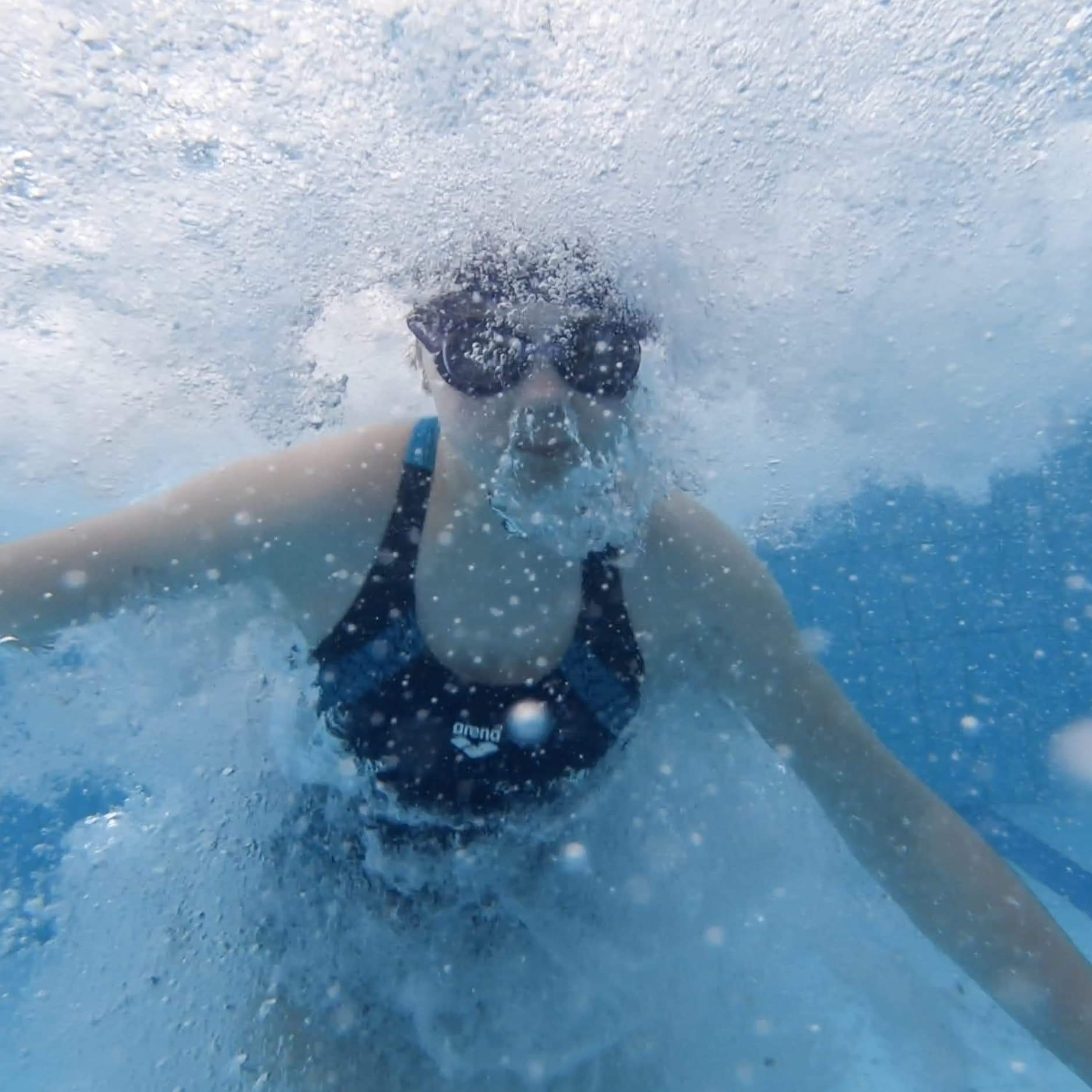 Osoba w okularach pływackich zanurzona w wodzie, widok podwodny, liczne pęcherzyki powietrza, strój kąpielowy marki Arena.