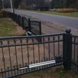 Automatyczna brama wjazdowa z czarnego metalu, widoczny mechanizm otwierający, w tle droga i zieleń.