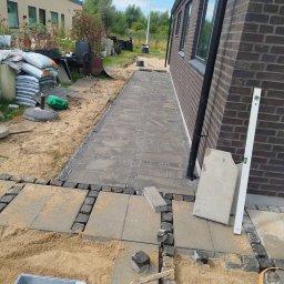 Układanie kostki brukowej: szara kostka ułożona wzdłuż ściany budynku, widoczne narzędzia i materiały budowlane, piasek. Etap prac wykończeniowych.