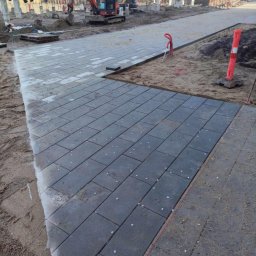 Częściowo ułożona, szara kostka brukowa na piasku, z widocznymi maszynami budowlanymi w tle. Widoczne białe elementy dystansowe między kostkami.
