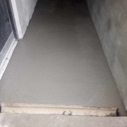 Wylana, świeża wylewka podłogowa w pomieszczeniu, wyrównana listwą. Widoczne ściany i fragment ramy drzwiowej. Szara kolorystyka, surowy stan wykończenia.