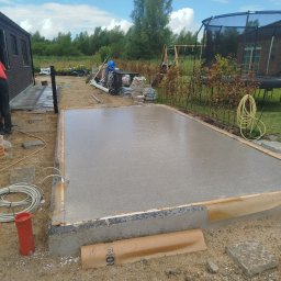 Wylana, świeża betonowa wylewka pod taras na zewnątrz domu, otoczona drewnianym szalunkiem. W tle widoczny ogród i elementy wyposażenia.