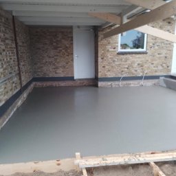 Ortyl - Wylana, świeża posadzka w garażu w Tarnobrzegu. Widoczne ceglane ściany, białe drzwi i okno. Konstrukcja dachu z drewnianych belek.