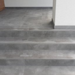 Ortyl - Wykończone płytkami schody wejściowe w odcieniach szarości, z widocznym fragmentem drzwi i elewacji budynku. Minimalistyczny design, czyste linie i stonowana kolorystyka.