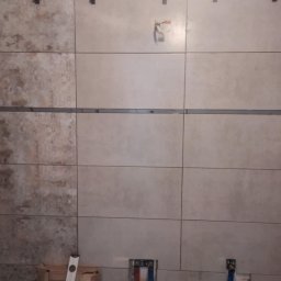 Ortyl - Ściana w trakcie wykańczania z beżowymi płytkami, widoczne elementy poziomowania i rury instalacji wodnej. Część płytek ma inny odcień.