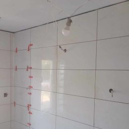 Ortyl - Białe płytki ścienne w trakcie montażu, z czerwonymi krzyżykami dystansowymi, surowa żarówka zwisająca z sufitu, widoczne otwory na instalacje.