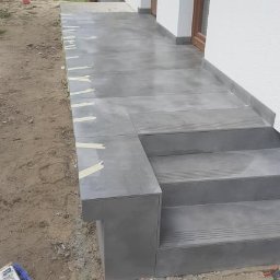 Ortyl - Wykończony taras z szarych płytek z widocznymi schodami i narzędziami glazurniczymi. Taśma malarska zabezpiecza krawędzie. Widok na dom z oknem.