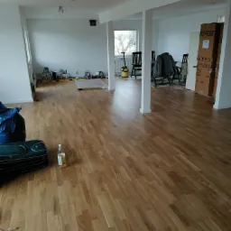 Jasne panele podłogowe w przestronnym pokoju po remoncie, z elementami wyposażenia i narzędziami w tle. Widoczna butelka i walizka na podłodze.