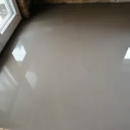 Świeżo wylana, gładka wylewka samopoziomująca w pomieszczeniu, widoczne refleksy światła, ściany w stanie surowym, okno z białymi ramami, folia ochronna na podłodze przy ścianie.