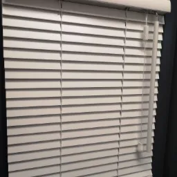 Zamknięte, białe żaluzje aluminiowe z widocznym mechanizmem sterującym, zamontowane w ciemnej ramie okiennej.