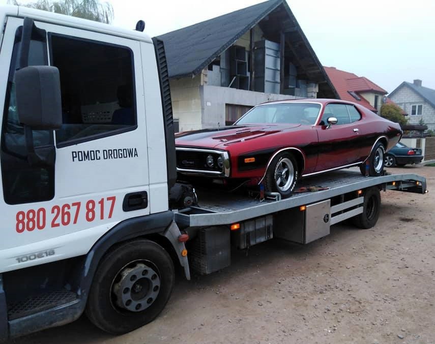 Czerwony, klasyczny samochód marki Dodge Charger z lat 70. na platformie lawety pomocy drogowej z napisem 'Pomoc Drogowa' i numerem telefonu, zaparkowanej na szutrowej drodze przed domem w trakcie...
