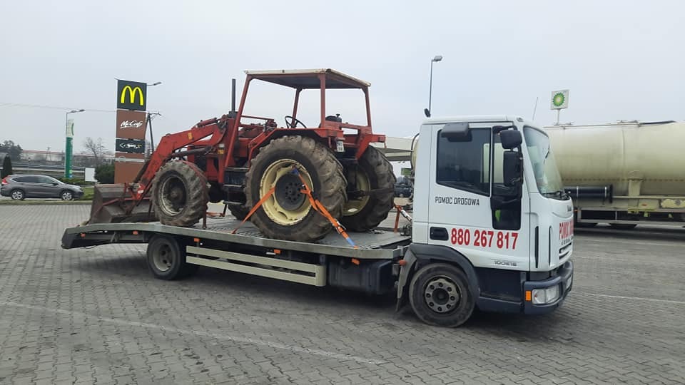 Laweta z czerwonym traktorem rolniczym z łyżką, zabezpieczonym pomarańczowymi pasami transportowymi, na tle stacji benzynowej i restauracji McDonald's w pochmurny dzień.