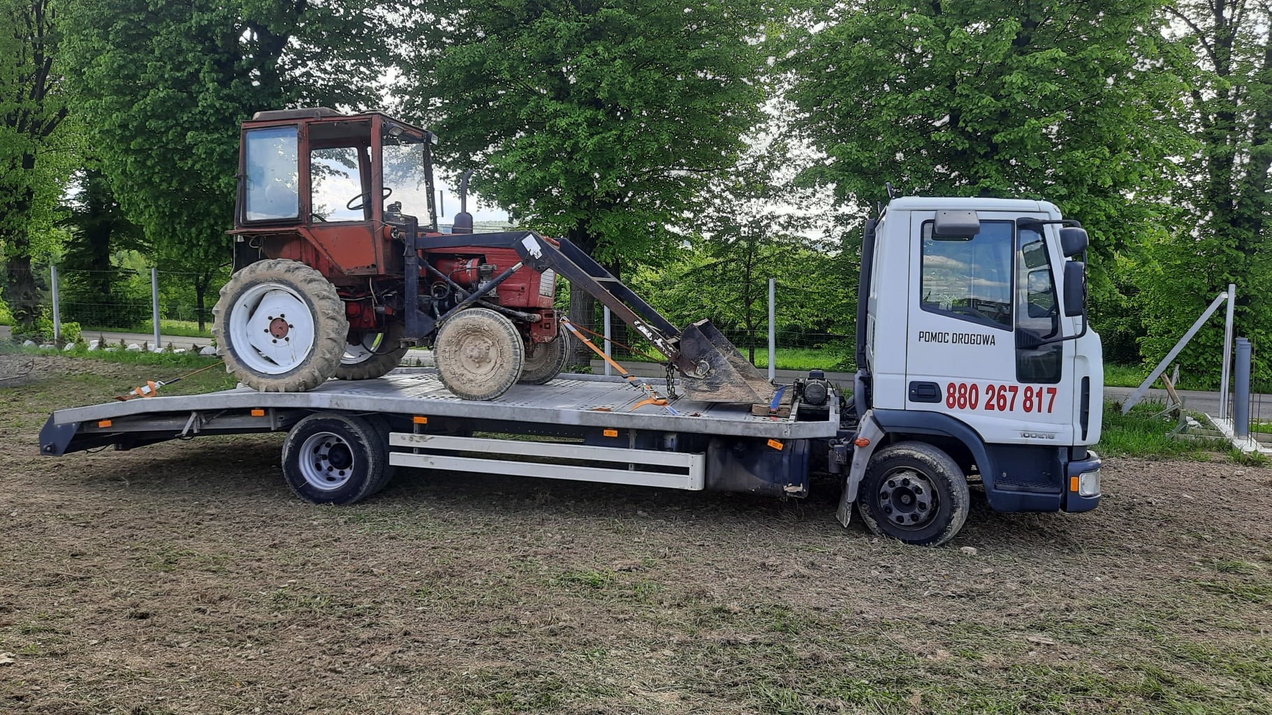 Czerwony traktor z łyżką załadowany na białą ciężarówkę z napisem 'Pomoc Drogowa', zaparkowaną na ziemi z widocznymi drzewami w tle.