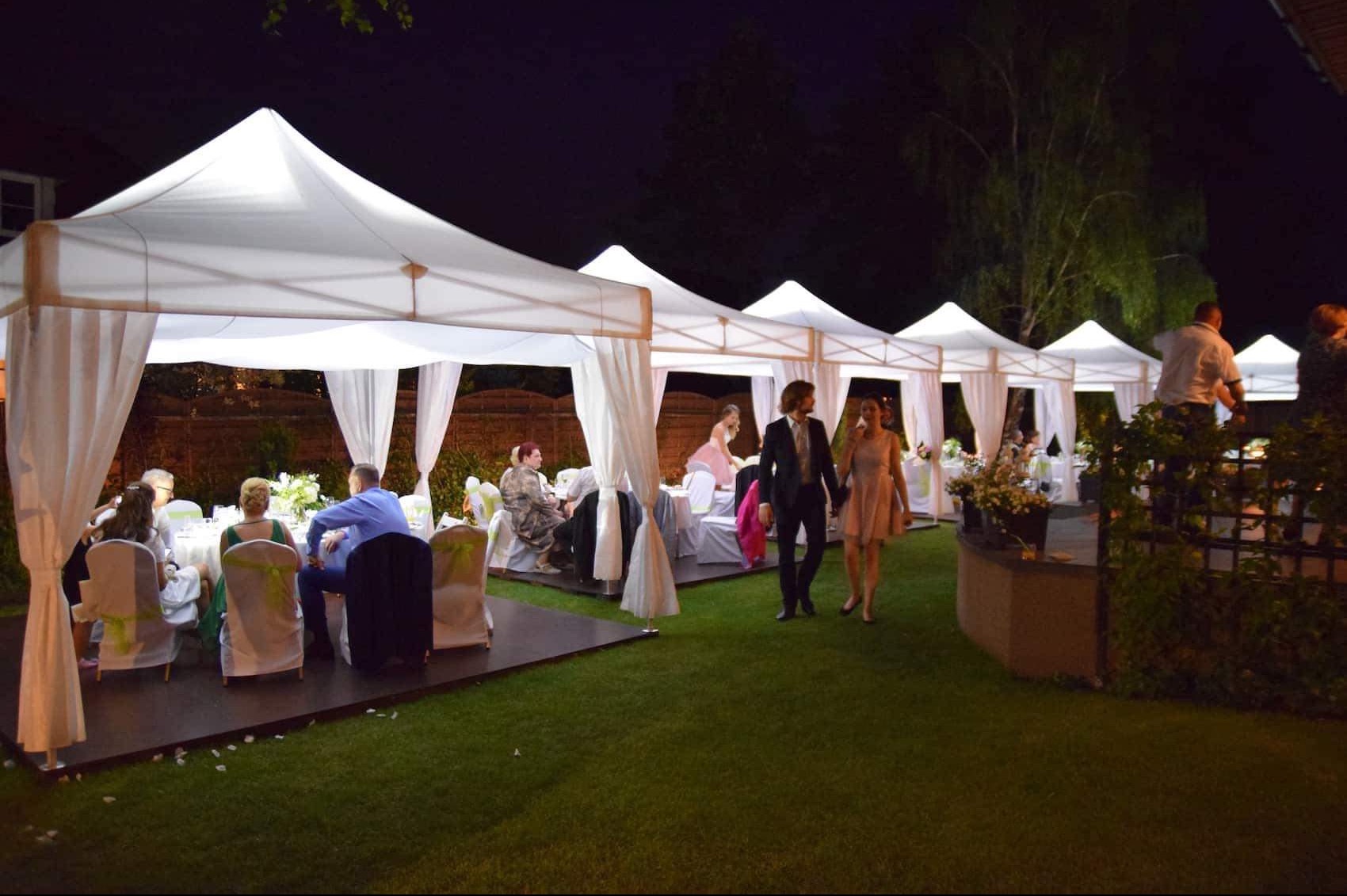 Uroczystość na zewnątrz w nocy: rzędy białych namiotów z zasłonami, pod którymi siedzą goście przy okrągłych stołach z kwiatami, para spaceruje po trawniku, a obsługa ustawia dekoracje.