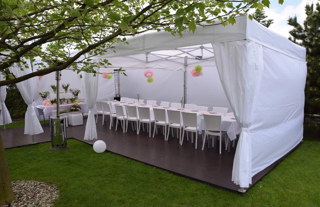 Biały namiot imprezowy z zasłonami, ustawiony na trawniku, z długim stołem i białymi krzesłami, udekorowany kwiatami i ozdobami w pastelowych kolorach, z widocznym grzejnikiem na gaz.