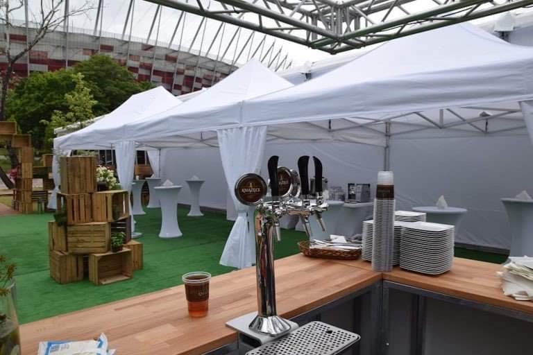 Białe namioty eventowe z zasłonami, zaaranżowane na zewnątrz z drewnianym barem z kranami do piwa, stosami talerzy i szklanek, z dekoracjami z drewnianych skrzynek i kwiatów, na tle stadionu.