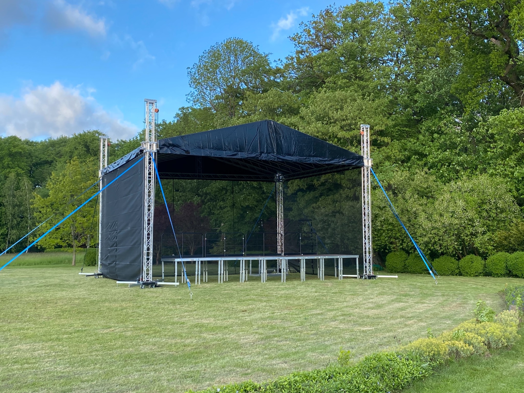 Czarna scena plenerowa z metalową konstrukcją na trawniku, zabezpieczona niebieskimi linami, w tle drzewa i błękitne niebo.