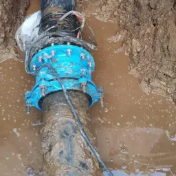 Montaż rury z niebieskim kołnierzem w wykopie wypełnionym wodą w Koszalinie, widoczne przewody i błotniste ściany wykopu.