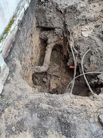 Głęboki wykop w ziemi z widocznymi rurami i kablami, fragment muru po lewej stronie, ziemia o różnej strukturze i wilgotności.
