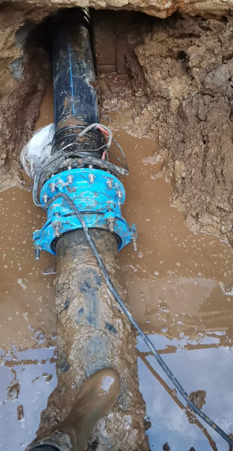 Montaż rury z niebieskim kołnierzem w wykopie wypełnionym wodą w Koszalinie, widoczne przewody i błotniste ściany wykopu.