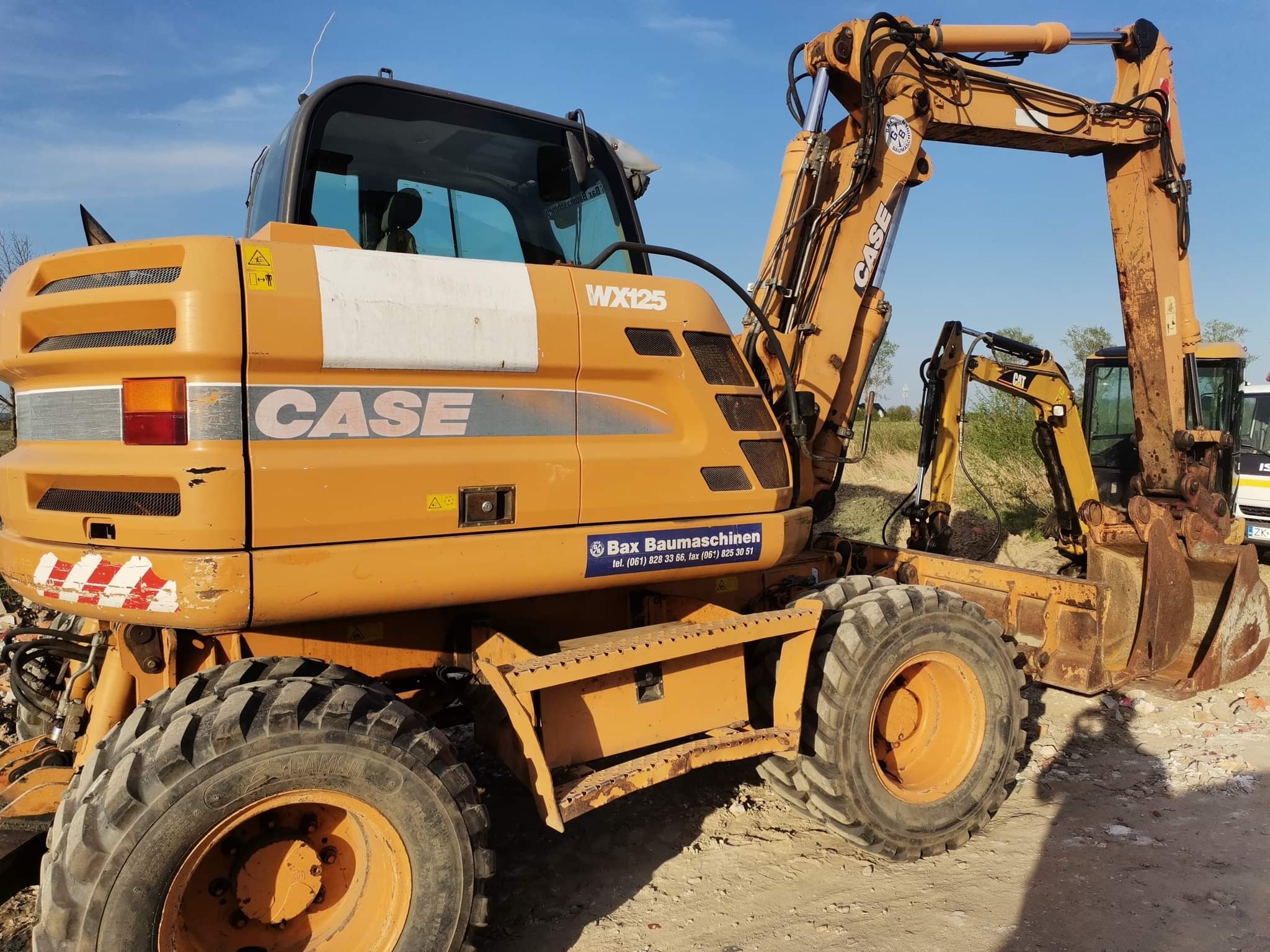 Żółta koparka kołowa Case WX125 z łyżką, widoczna z boku, na placu budowy, z widocznymi śladami użytkowania i logo firmy Bax Baumaschinen.