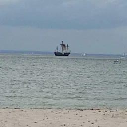 Widok na morze z piaszczystej plaży, na horyzoncie żaglowiec stylizowany na galeon.